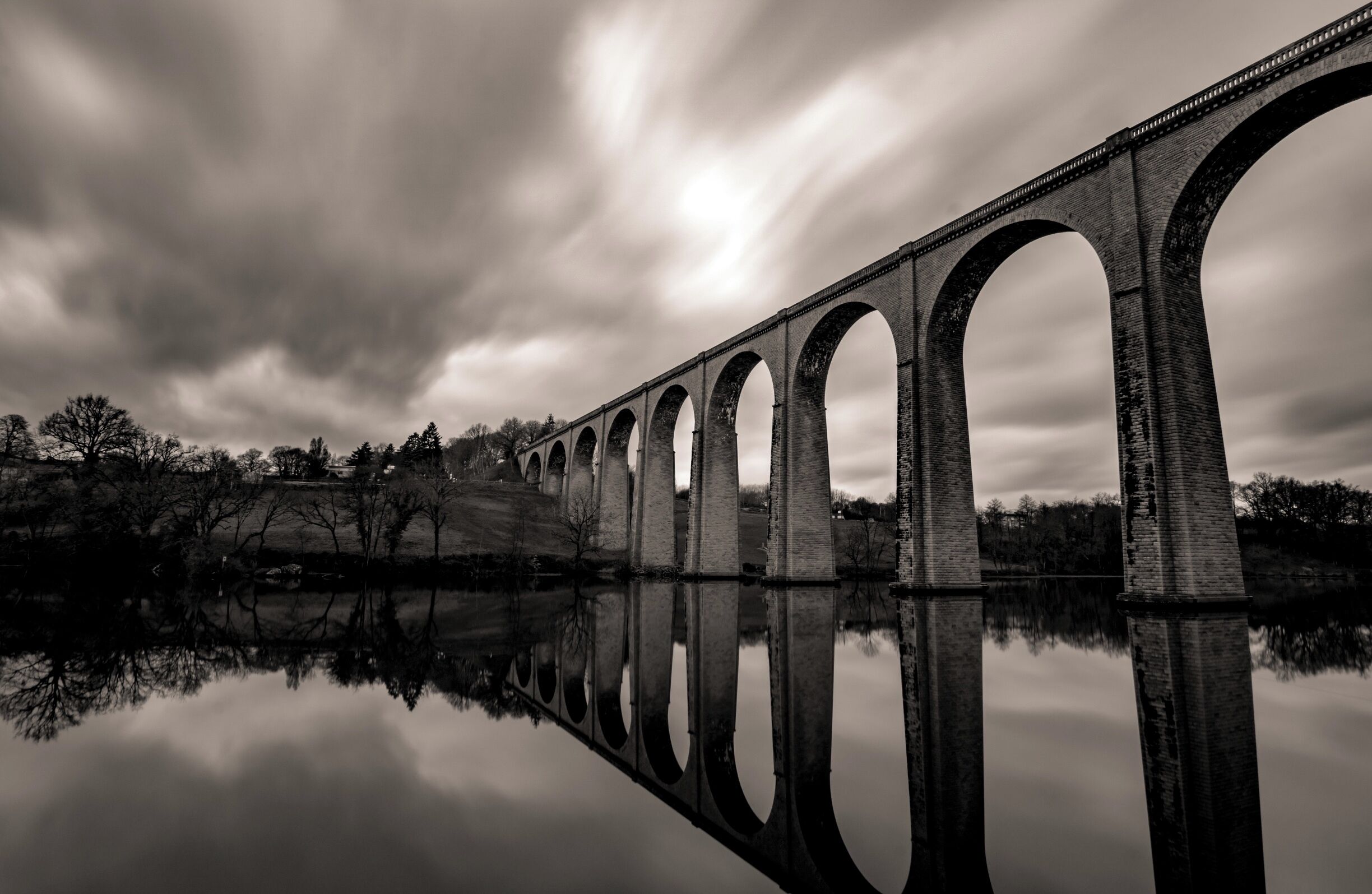 A long bridge with multiple arches spanning a calm body of water, reflecting the structure.