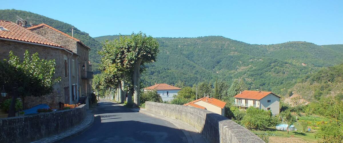 A narrow road flanked by stone walls and buildings with red-tiled roofs, leading through a hilly landscape.