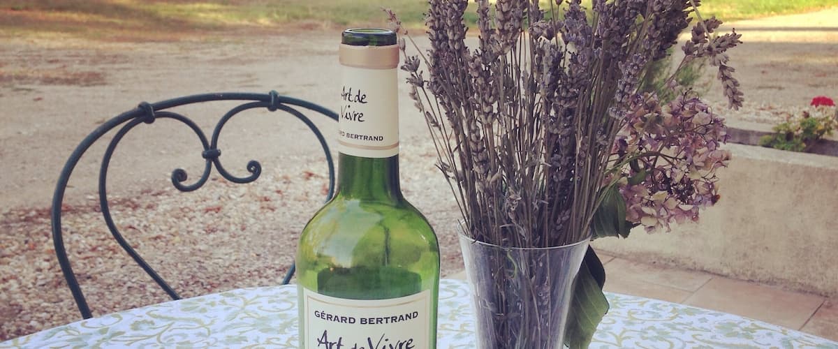 A bottle of Art de Vivre Merlot wine, a glass of red wine, and a vase with lavender on an outdoor table.