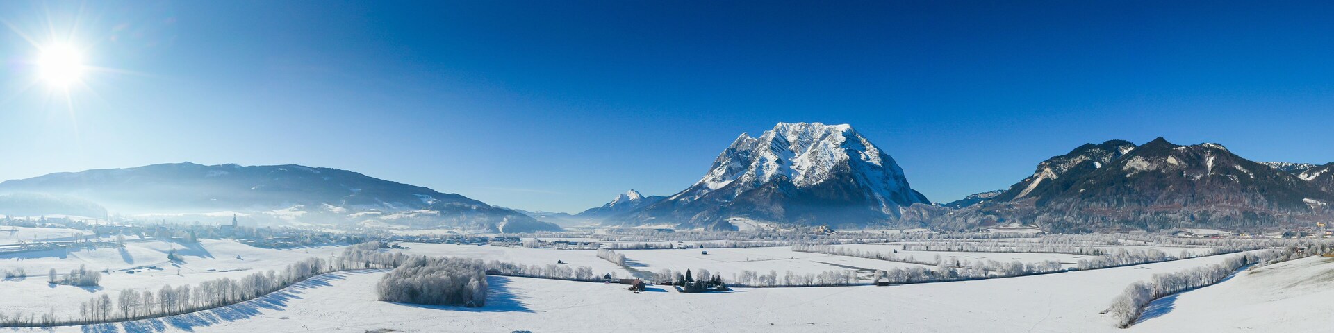 Paisagem coberta de neve com um pico de montanha imponente, um rio e árvores dispersas.