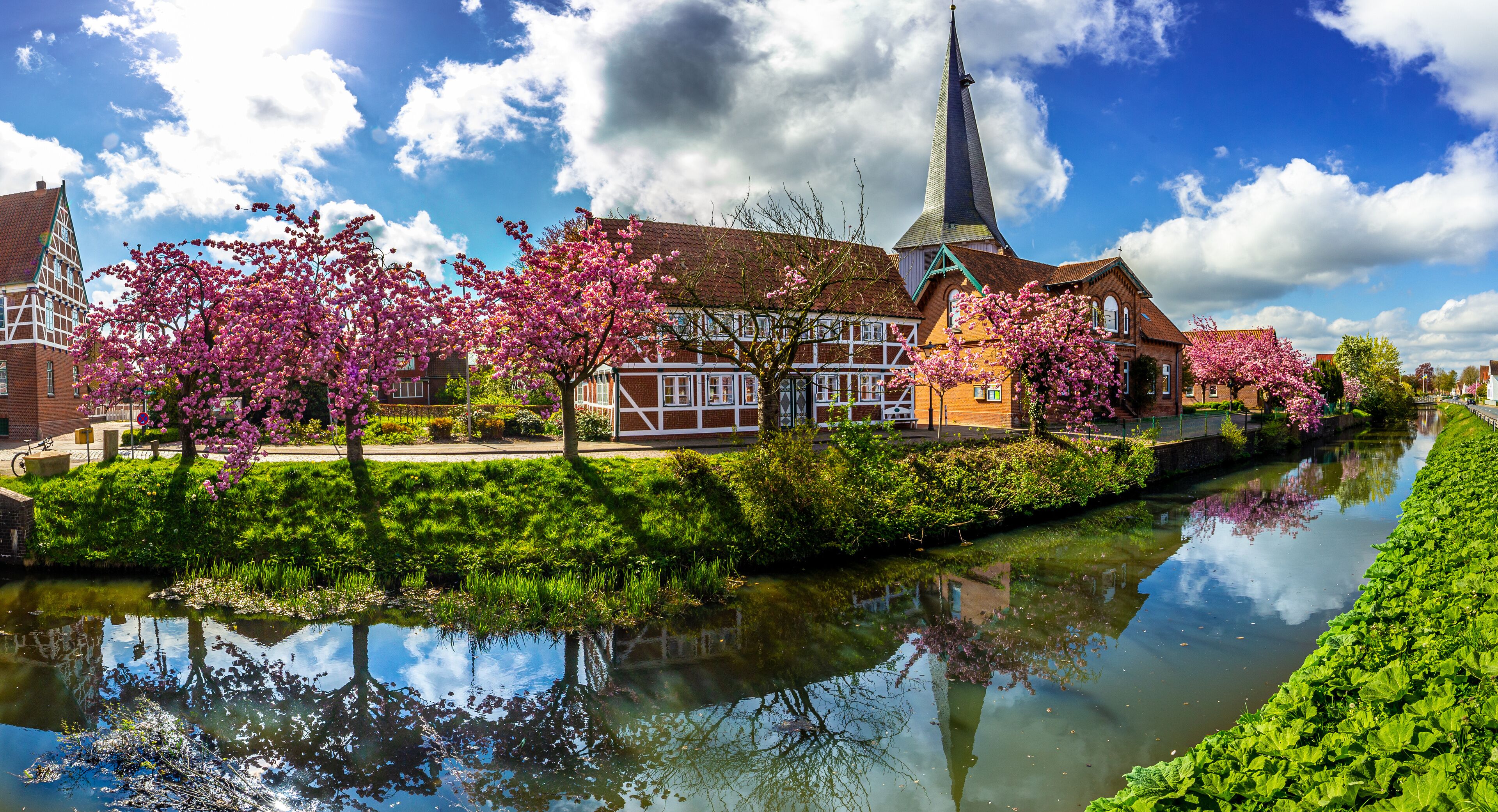 A canal with reflections, a church with a tall steeple, and blooming cherry trees.
