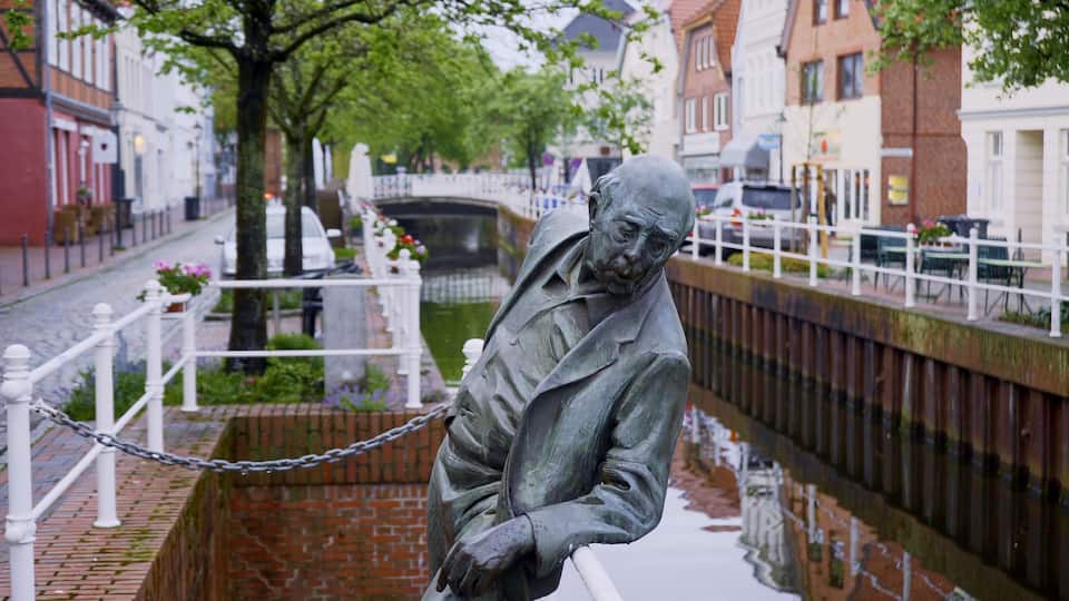 A statue of a man in a suit by a canal with a bridge in the background.