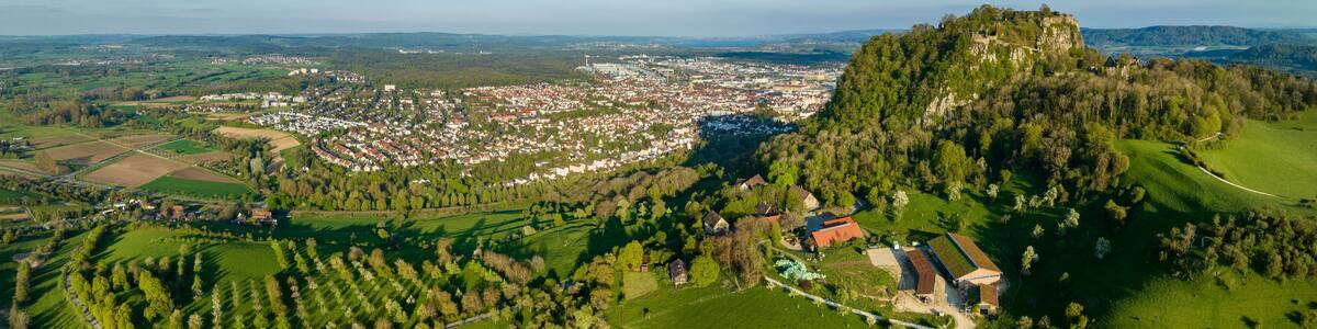 Luftbild-Panorama vom Vulkankegel Hohentwiel mit der Burgruine von der Abendsonne angestrahlt, dahinter die Stadt Singen am Hohentwiel