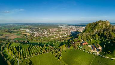 Luftbild-Panorama vom Vulkankegel Hohentwiel mit der Burgruine von der Abendsonne angestrahlt, dahinter die Stadt Singen am Hohentwiel
