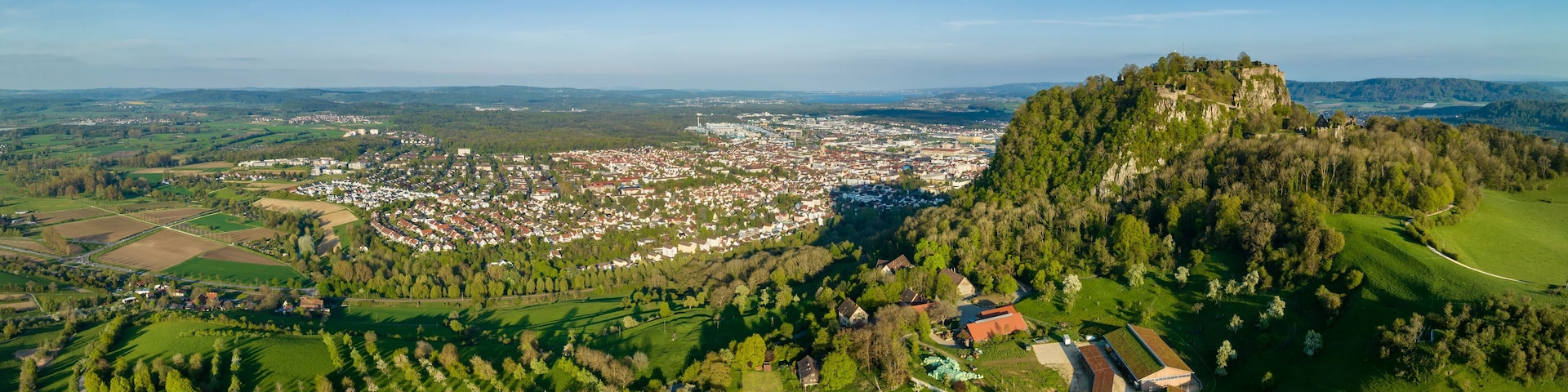 Luftbild-Panorama vom Vulkankegel Hohentwiel mit der Burgruine von der Abendsonne angestrahlt, dahinter die Stadt Singen am Hohentwiel
