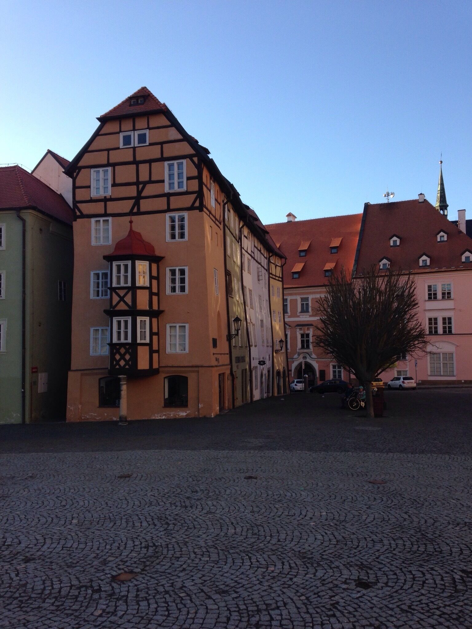 A cobblestone square with traditional half-timbered buildings, a tree, and a clear sky.
