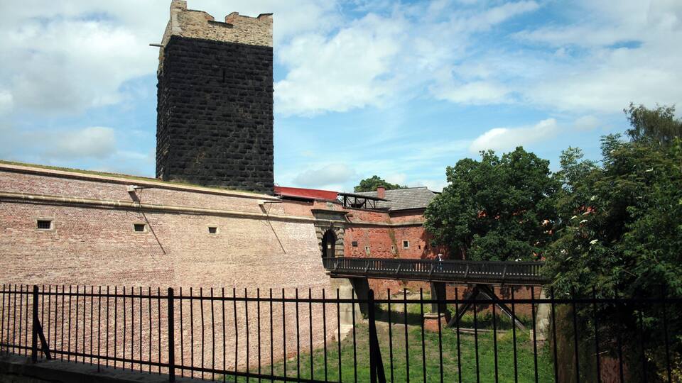 A historic brick wall with a tall tower, a metal fence, and a tree in the foreground.