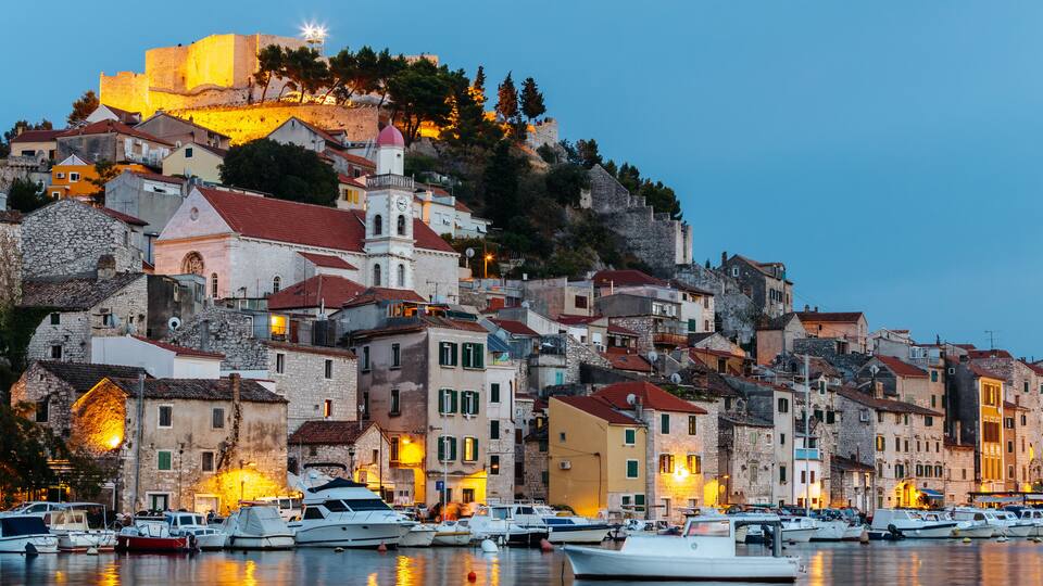 A coastal town with historic stone buildings, a prominent castle on a hill, and a marina with boats.
