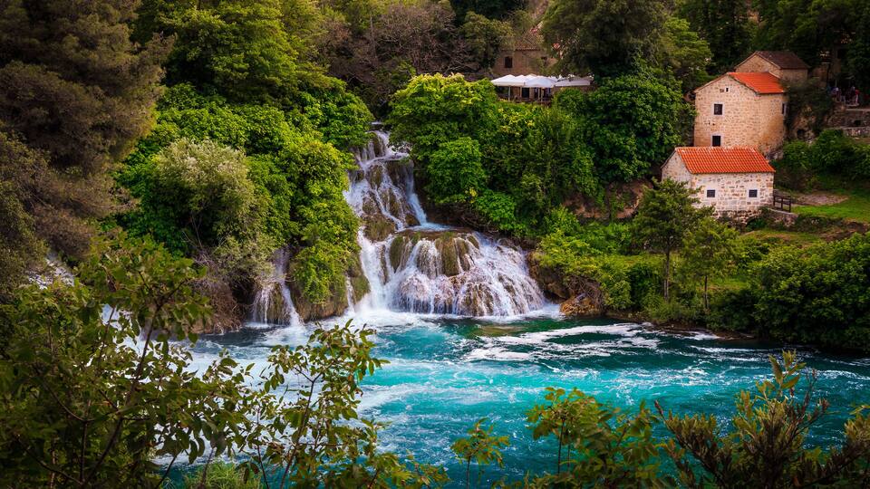 A waterfall cascading into a turquoise river surrounded by lush greenery and stone buildings.