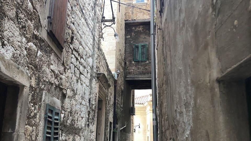 Un estrecho callejón adoquinado flanqueado por edificios antiguos de piedra con contraventanas de madera y un farol.
