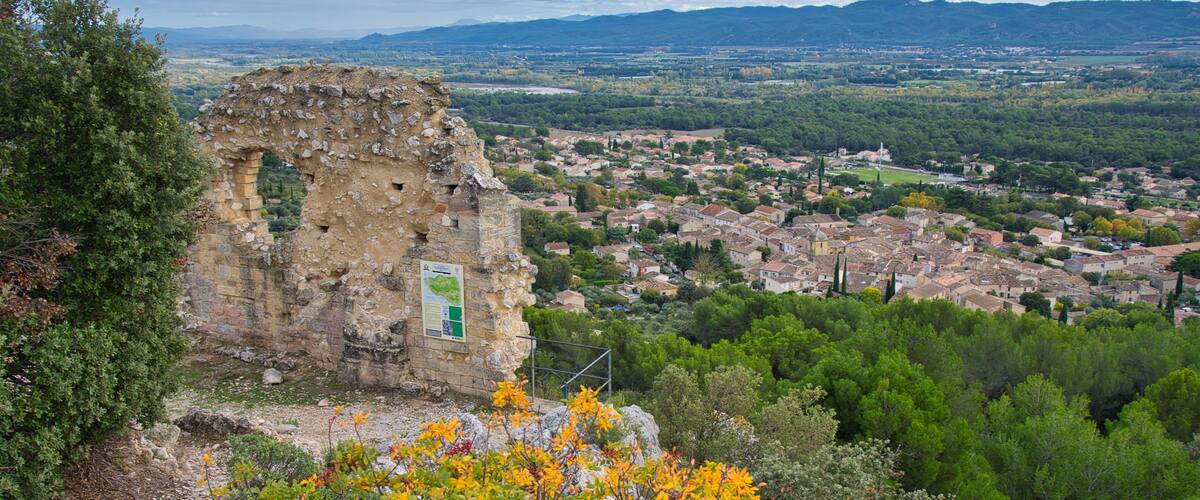 A historic stone ruin with a green sign, surrounded by lush greenery and overlooking a town with houses and mountains in the background.