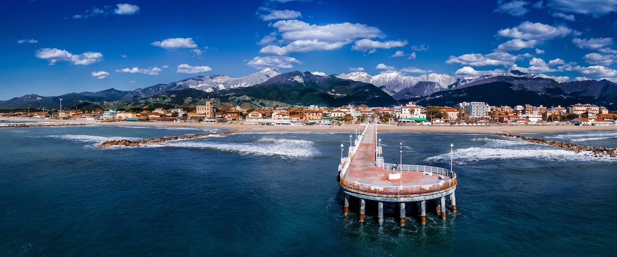 A coastal town with a pier extending into the sea, mountains in the background, and a clear blue sky.