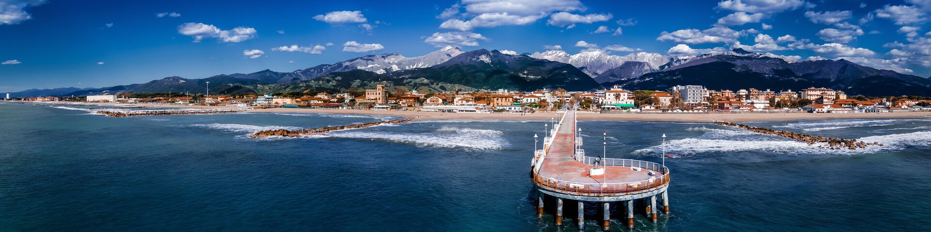 A coastal town with a pier extending into the sea, mountains in the background, and a clear blue sky.