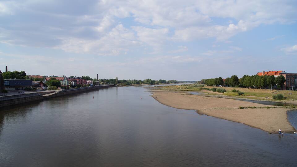 A river with a sandy bank, buildings, and trees in the background.