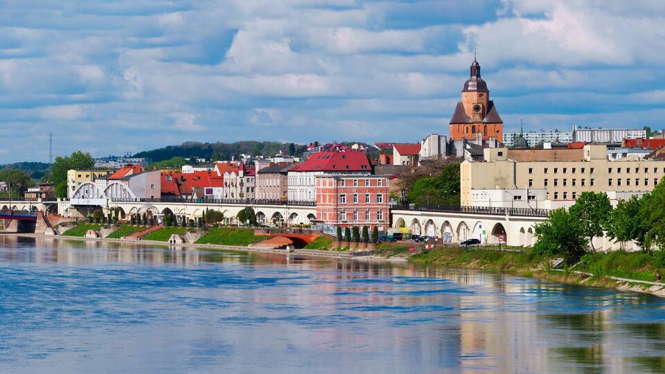 A river with a bridge, a prominent church tower, and a mix of historic and modern buildings along the riverbank.