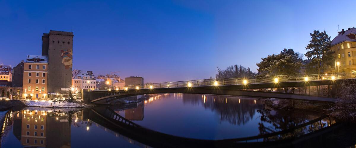 A bridge over a calm river reflecting buildings and trees.