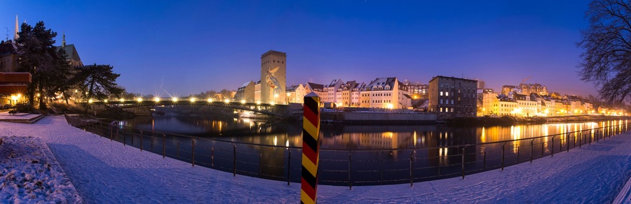 Ein mit Schnee bedeckter Fluss mit einer Brücke und beleuchteten Gebäuden im Hintergrund.