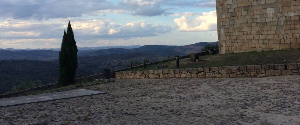 A stone tower with a crenellated top, a cobblestone pathway, and a distant view of a mountain range under a partly cloudy sky.