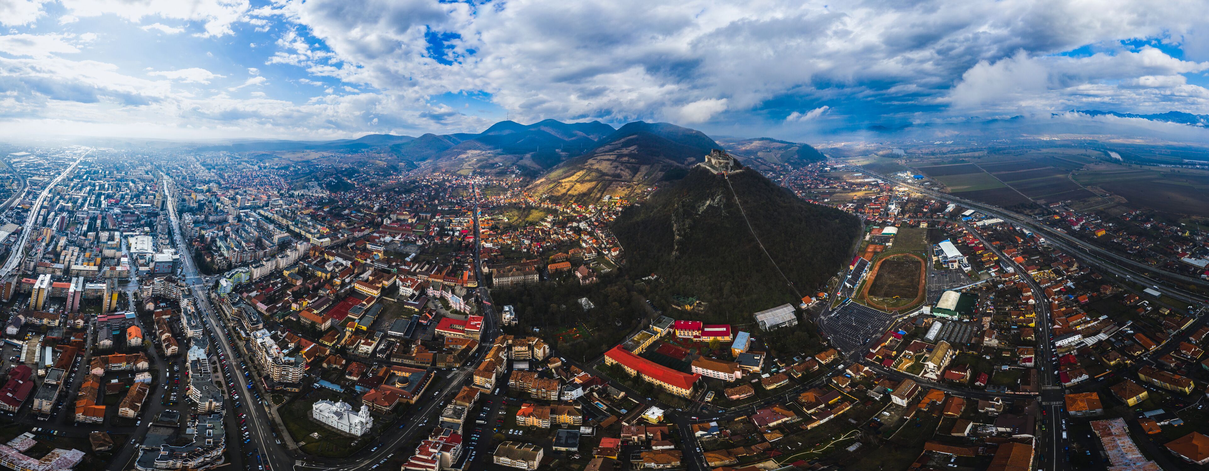 An aerial view of a city with a prominent mountain in the background, a large stadium, and a dense urban area with many buildings and roads.