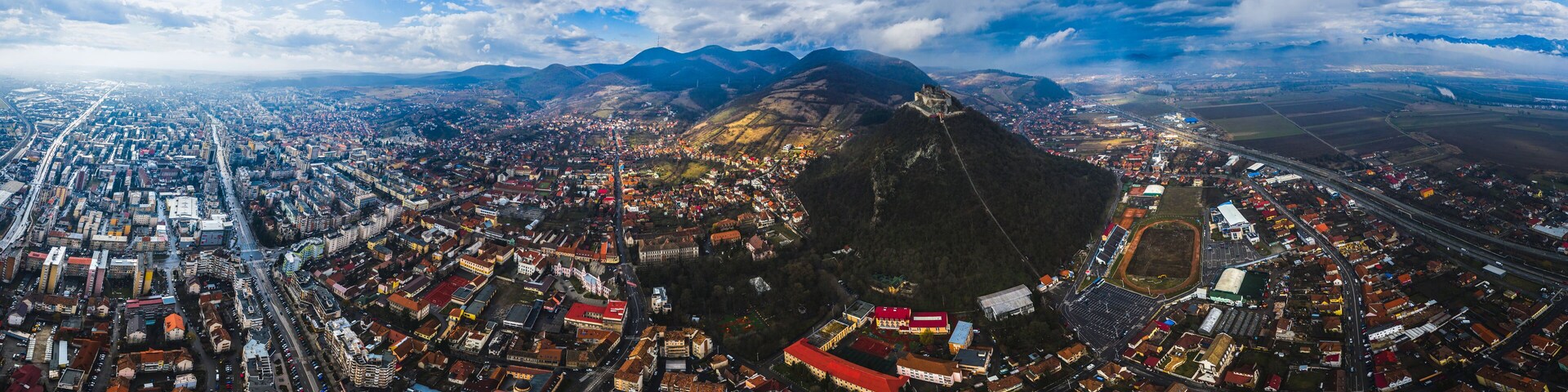 An aerial view of a city with a prominent mountain in the background, a large stadium, and a dense urban area with many buildings and roads.