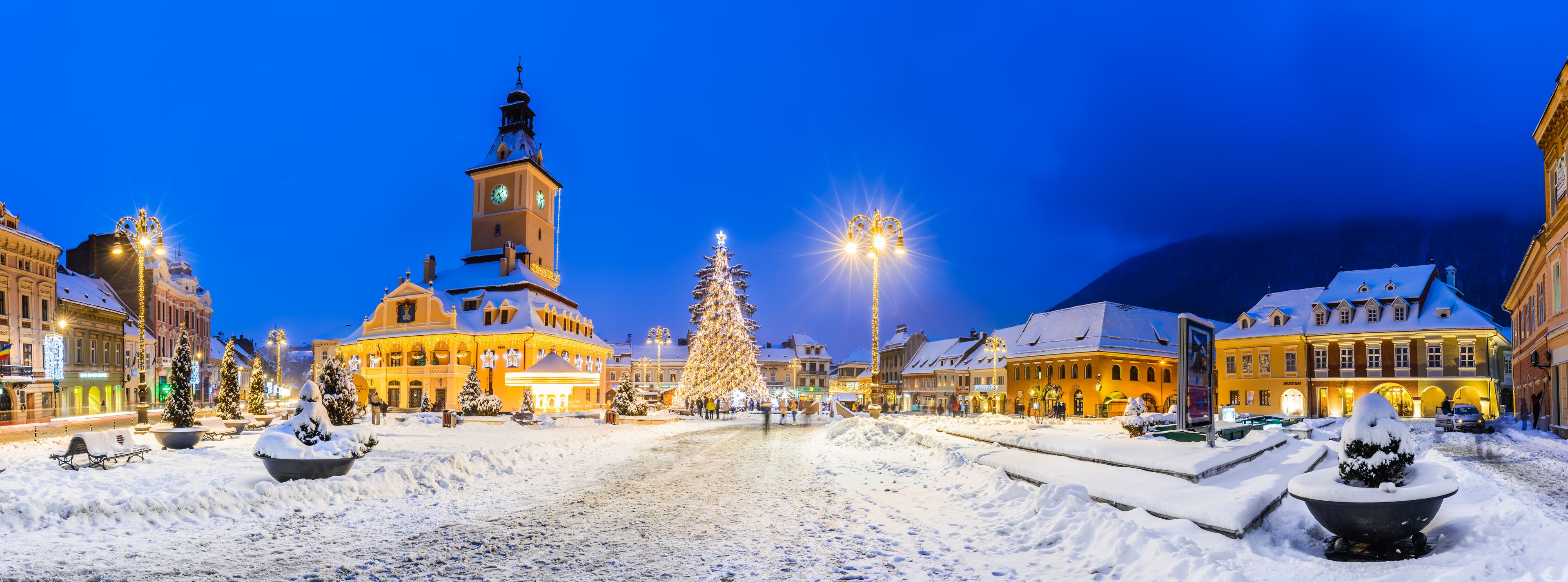 A snow-covered square with a prominent clock tower, illuminated buildings, and a large decorated Christmas tree.