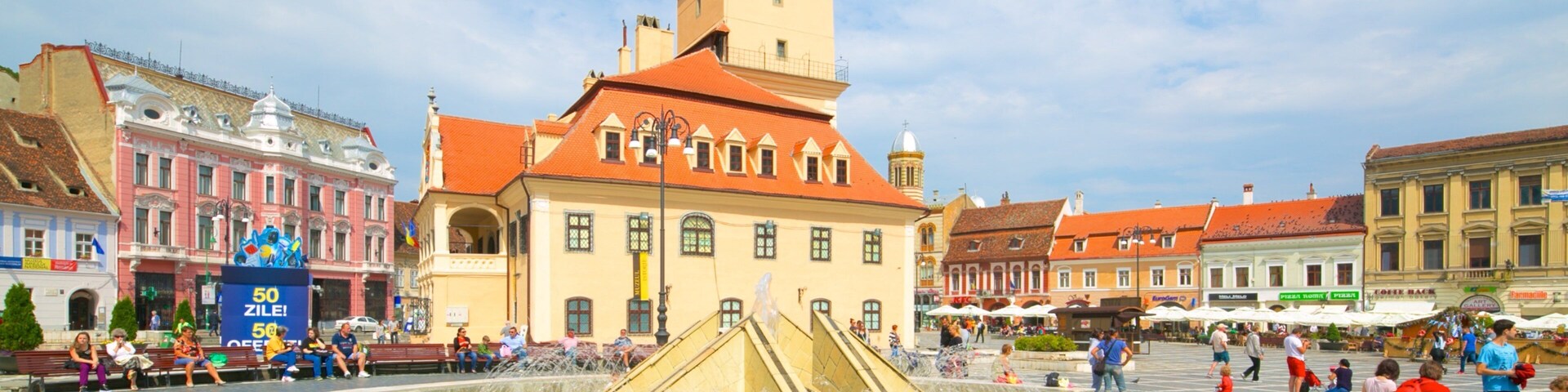 A historic square with a central fountain, a clock tower, and surrounding buildings.