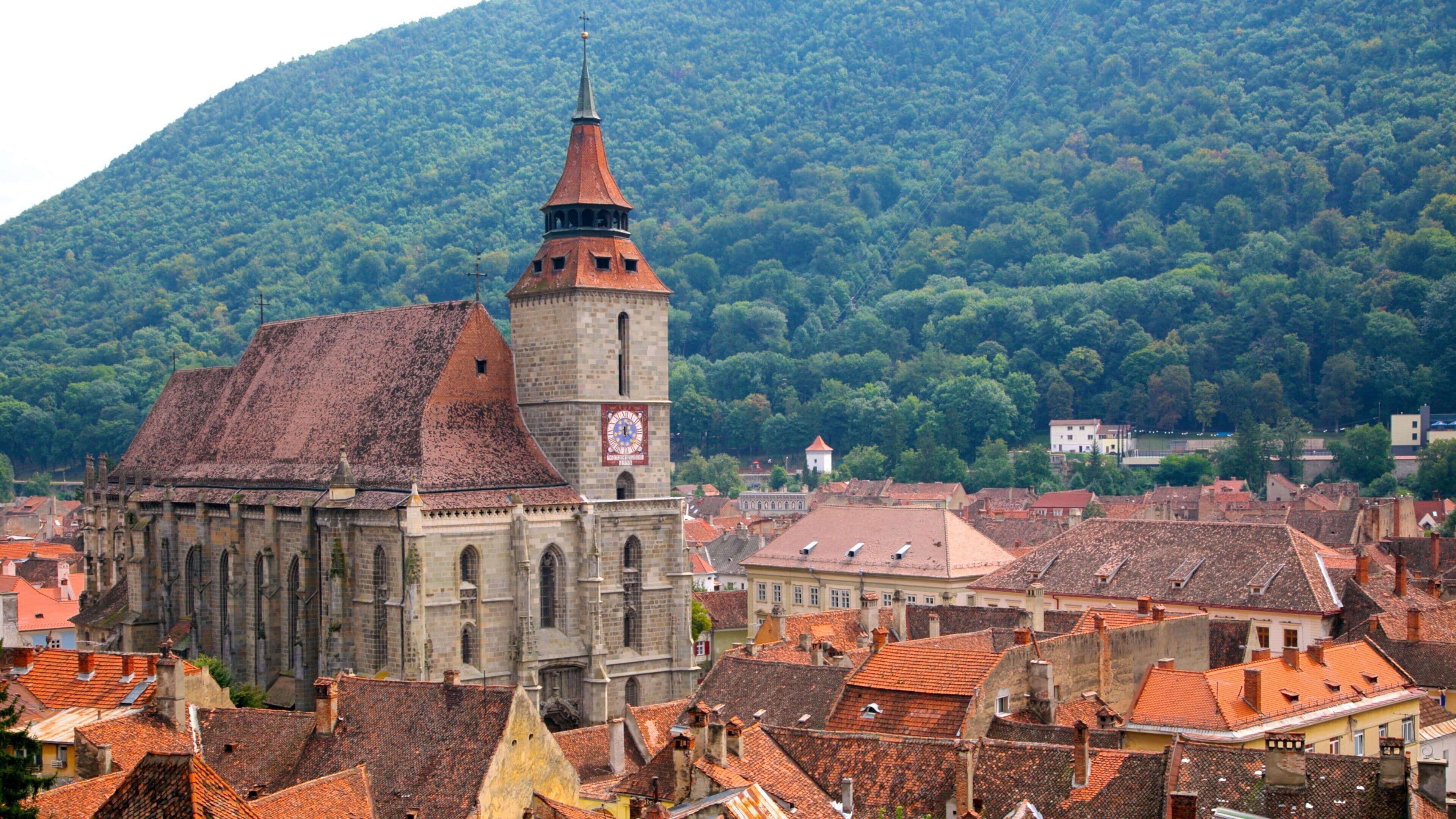 A historic church with a prominent tower and a clock, surrounded by traditional buildings and a forested hill in the background.