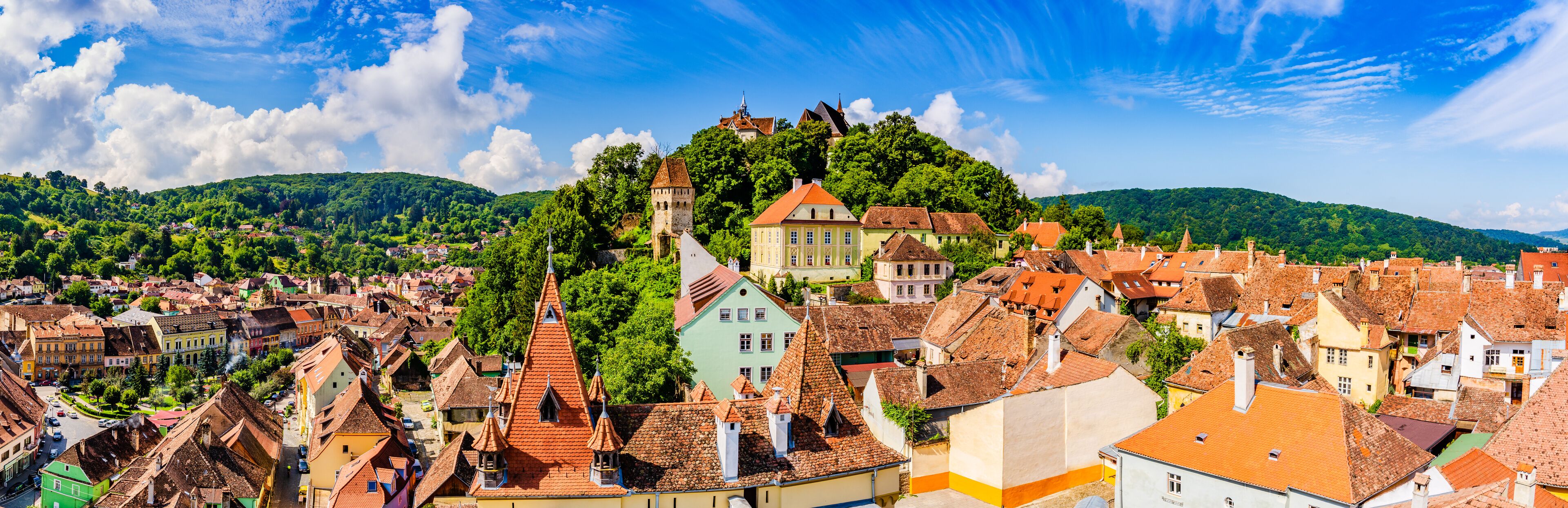 A town with colorful buildings, a prominent tower, and lush green hills in the background.
