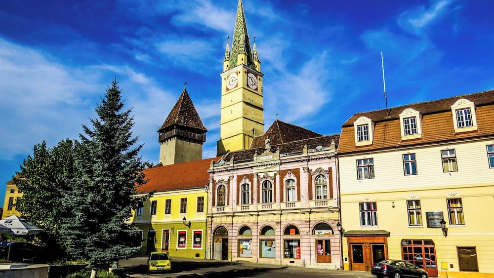 Ein historischer Stadtplatz mit einem markanten Glockenturm, bunten Gebäuden und blauem Himmel.