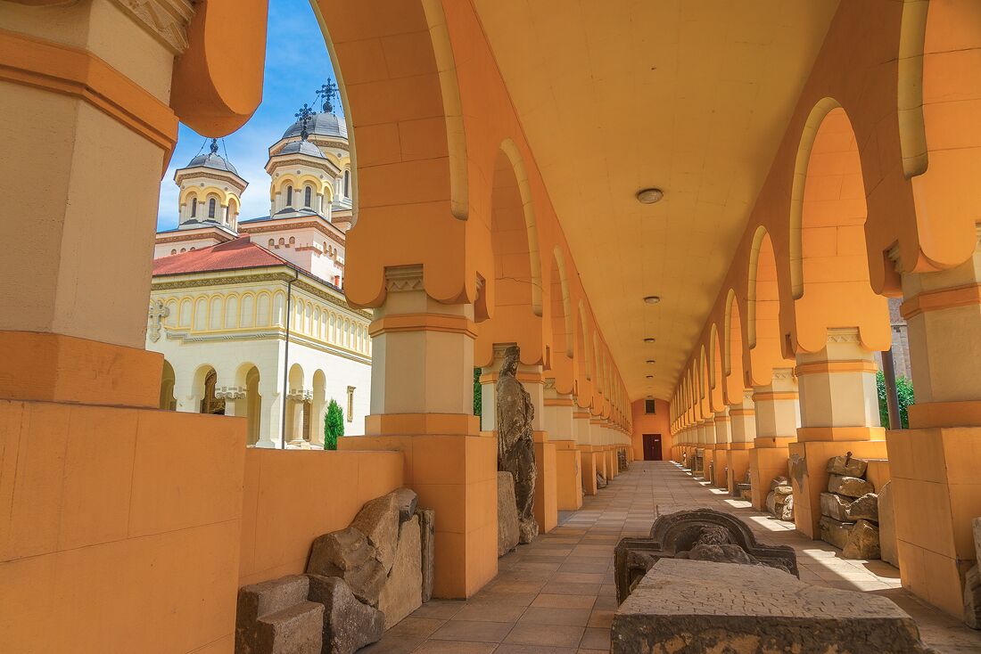 A corridor with arches and a statue, leading to a building with domed towers.