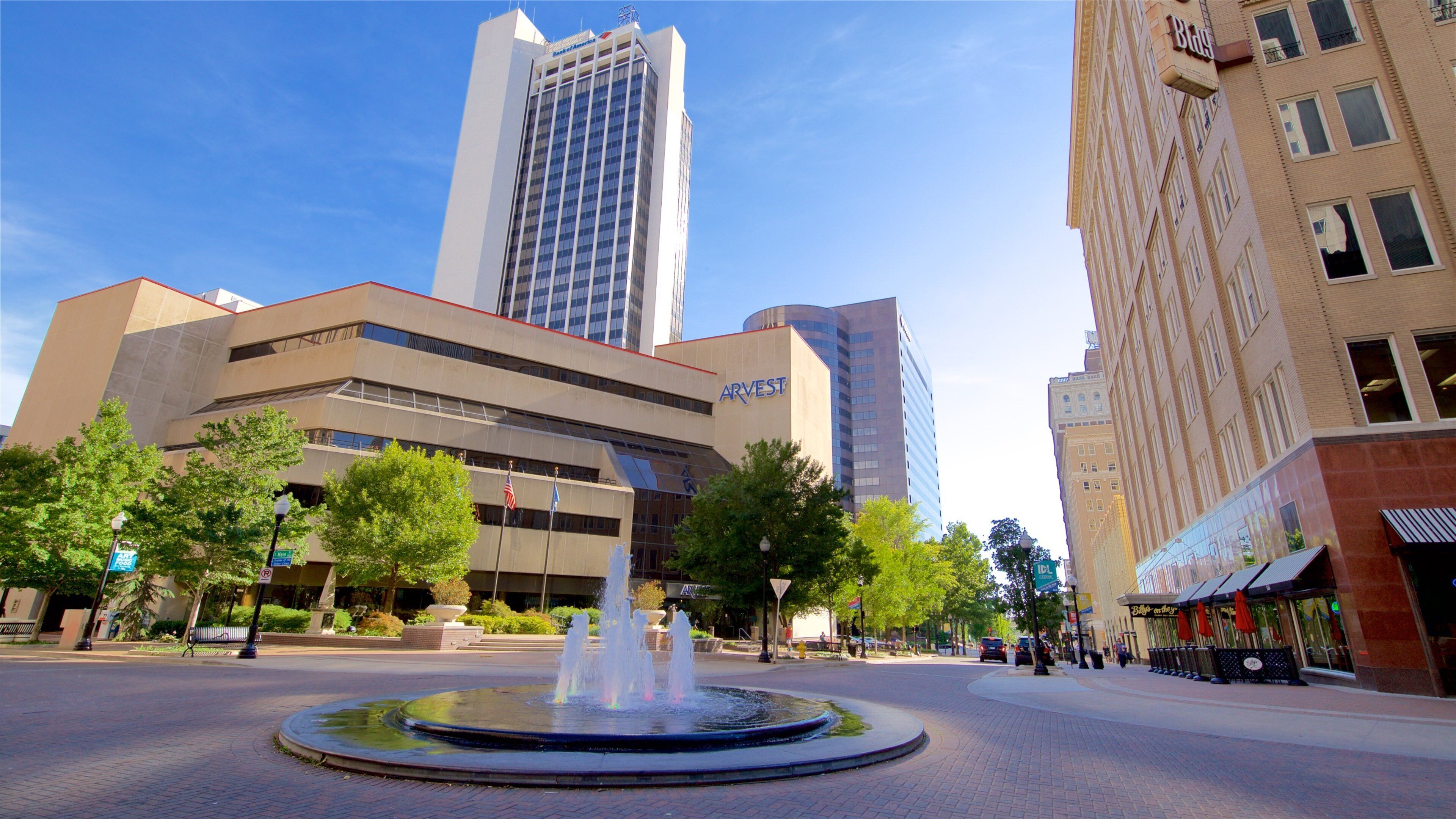 A city square with a fountain, surrounded by modern buildings including a tall skyscraper and a bank.