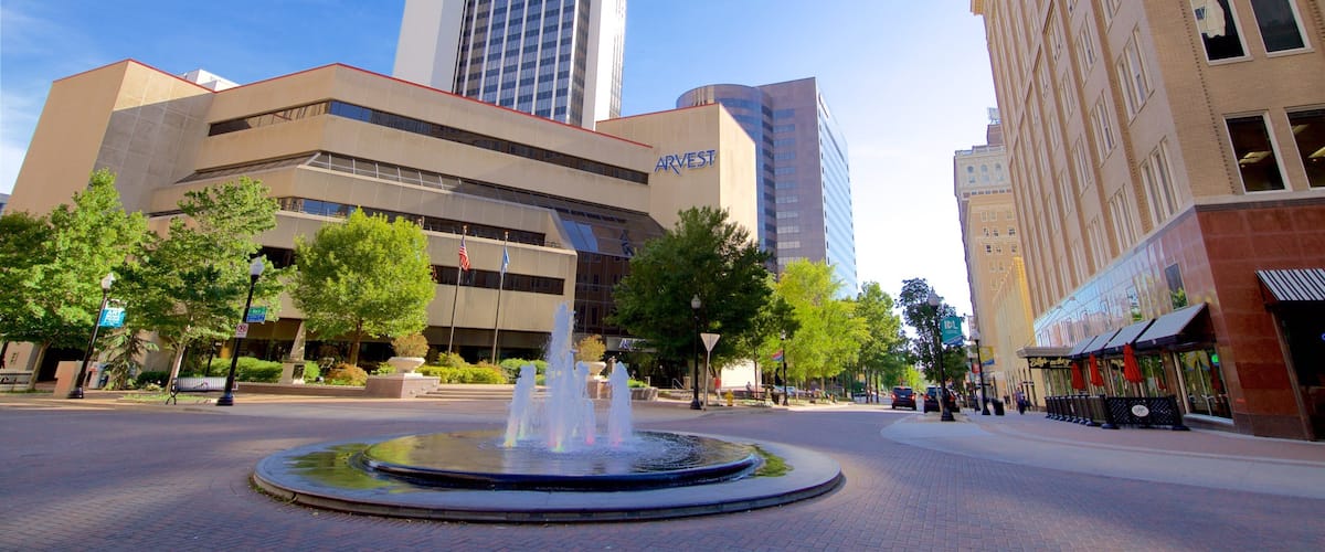 A city square with a fountain, surrounded by modern buildings including a tall skyscraper and a bank.
