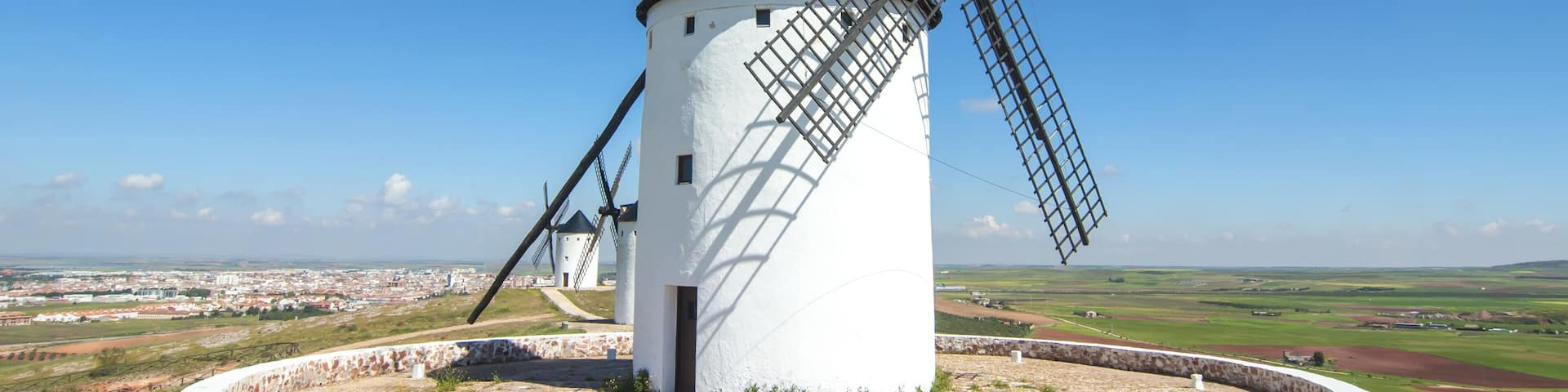 A white windmill with a conical roof and large sails, surrounded by a stone base and a circular stone wall, set against a clear blue sky and a distant cityscape.