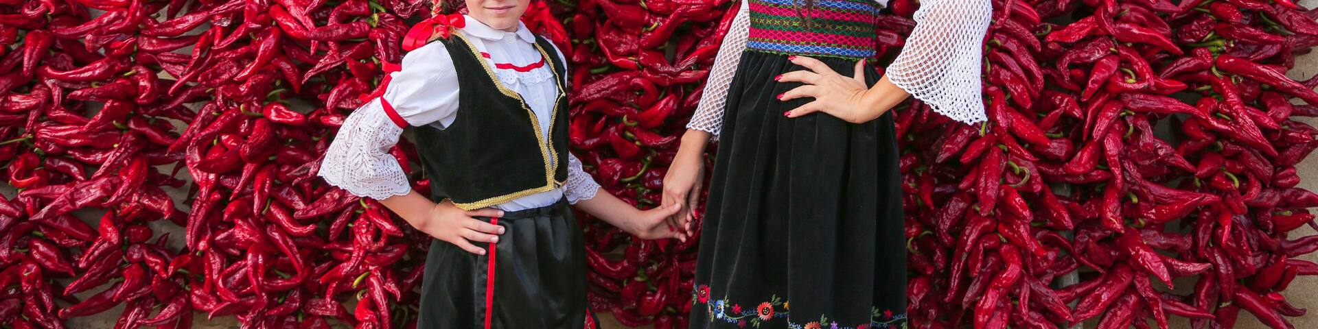 Girls dressed on traditional Serbian Balkan clothing, national folk costume. Posing near of lot red paprika peppers hanging on the wall of house. .