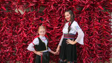 Girls dressed on traditional Serbian Balkan clothing, national folk costume. Posing near of lot red paprika peppers hanging on the wall of house. .