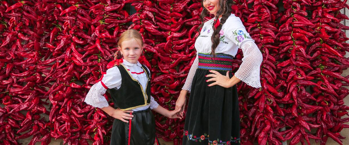 Two people in traditional attire standing in front of a wall of red chili peppers.