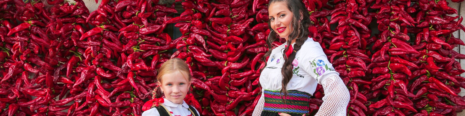 Two people in traditional attire standing in front of a wall of red chili peppers.