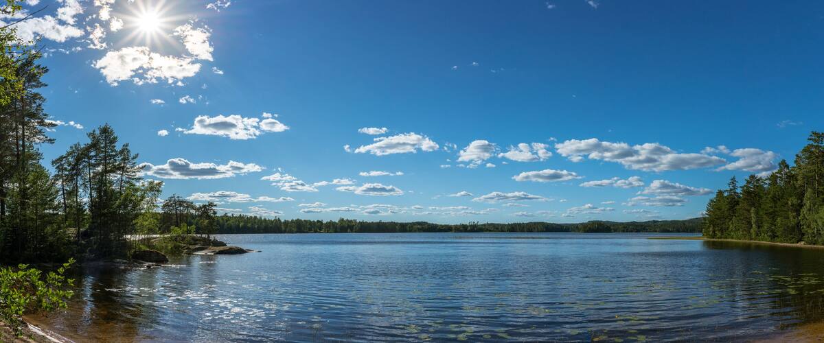A calm lake surrounded by dense forests and a clear blue sky with scattered clouds.