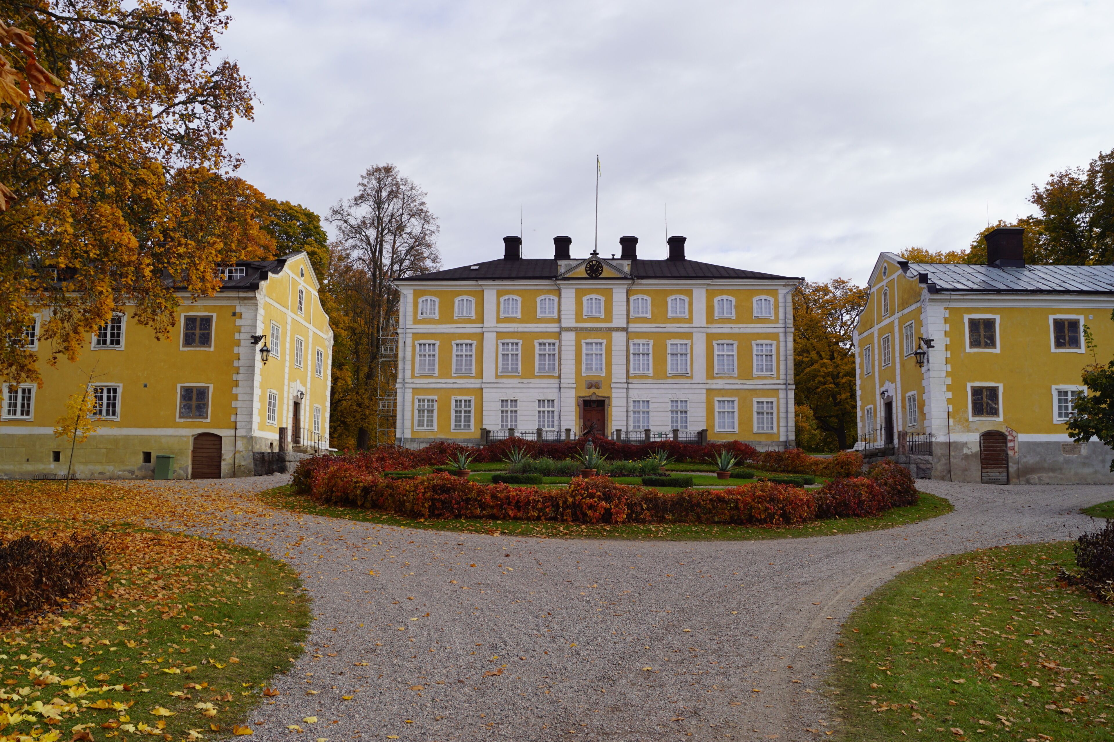 A large yellow building with a symmetrical facade, surrounded by a gravel path and a well-maintained garden with autumn foliage.