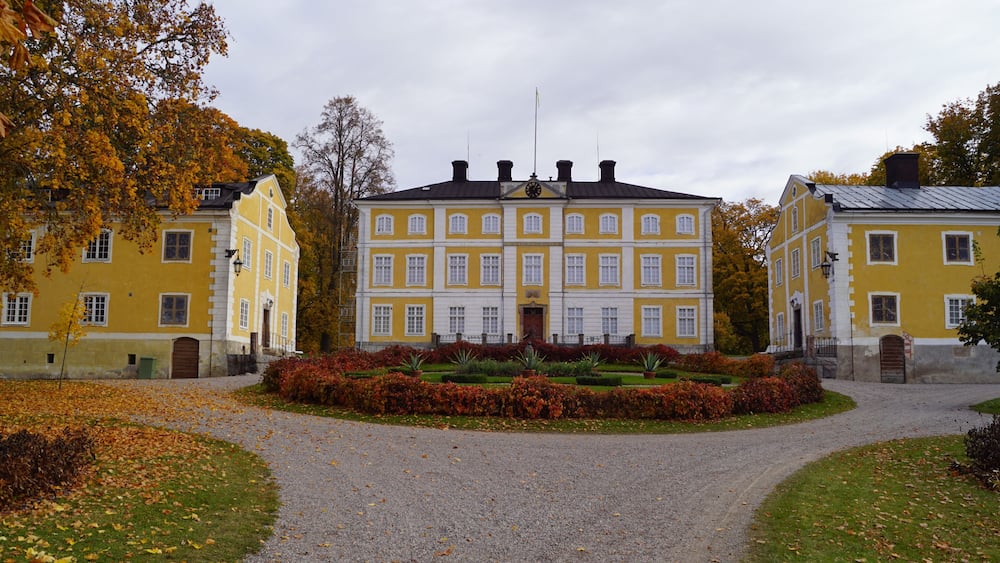 A large yellow building with a symmetrical facade, surrounded by a gravel path and a well-maintained garden with autumn foliage.