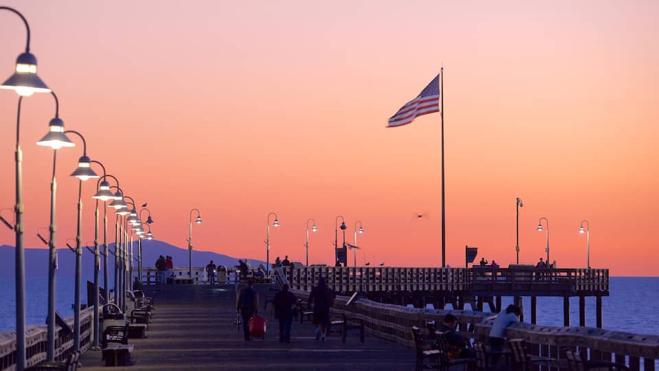 A pier at sunset with a flag, street lamps, and people walking.