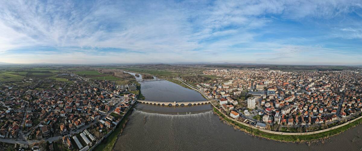 A panoramic view of a town with a prominent bridge over a river, surrounded by fields and distant hills.
