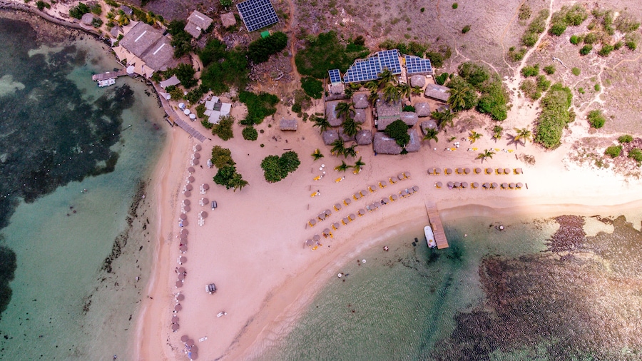 Vista aérea de una playa con filas de tumbonas, un muelle y paneles solares.