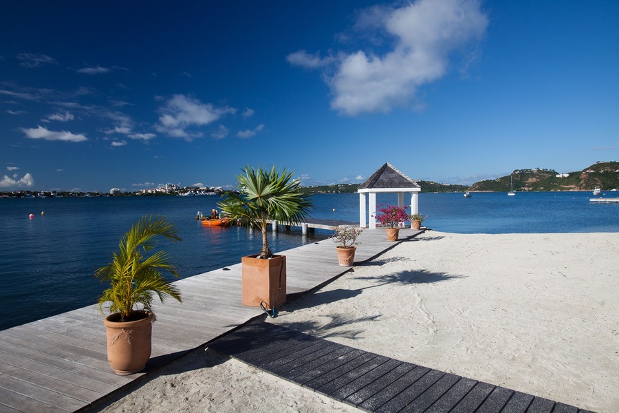 A wooden pier leading to a white gazebo by a calm sea with boats and a distant shoreline.