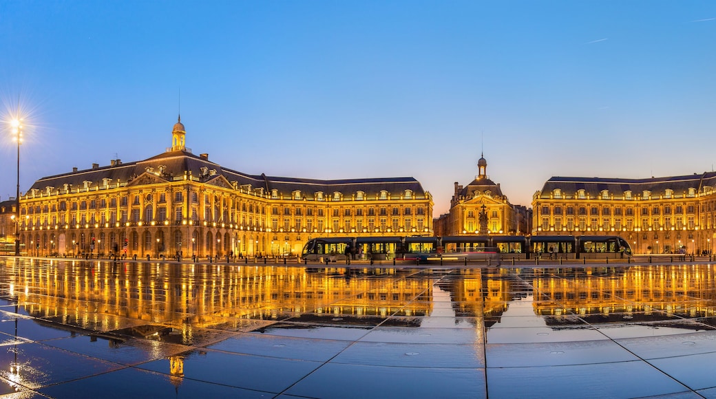 Un imposant bâtiment illuminé, doté d’un dôme central et de deux ailes symétriques, dont le reflet se mire sur une place humide.