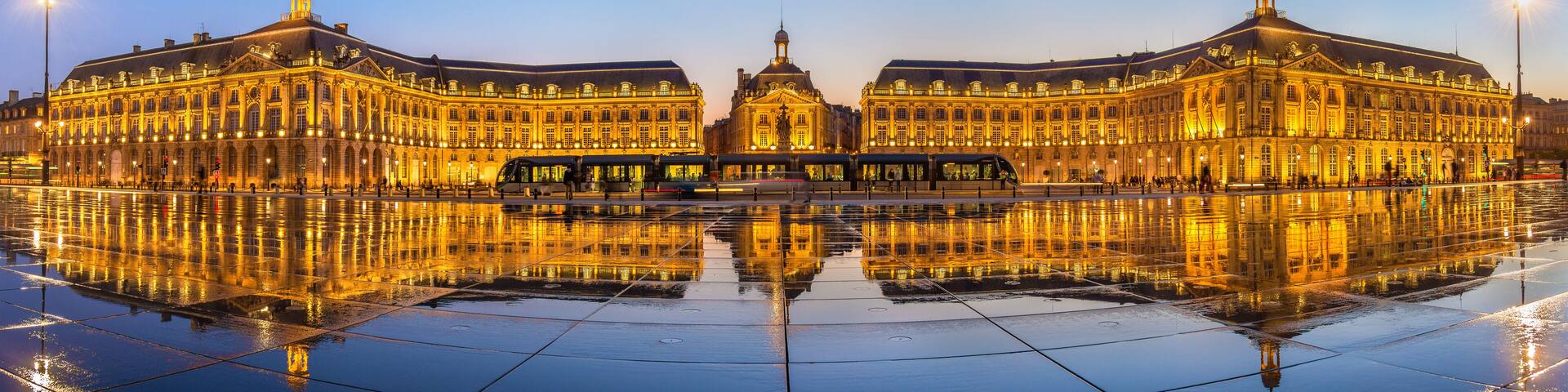 A grand illuminated building with a central dome and two symmetrical wings, reflecting on a wet plaza.