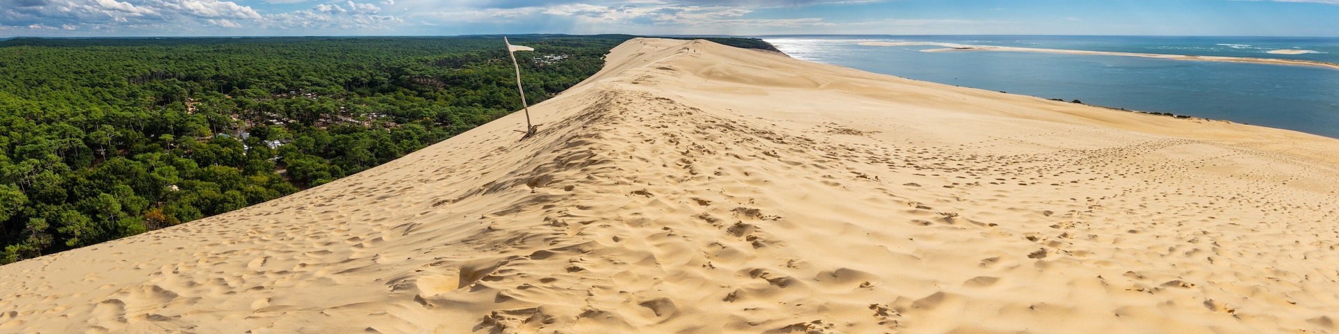 Eine Dünenlandschaft mit einem Flaggendenkmal, einem bewaldeten Gebiet im Hintergrund und einem Gewässer in der Ferne.