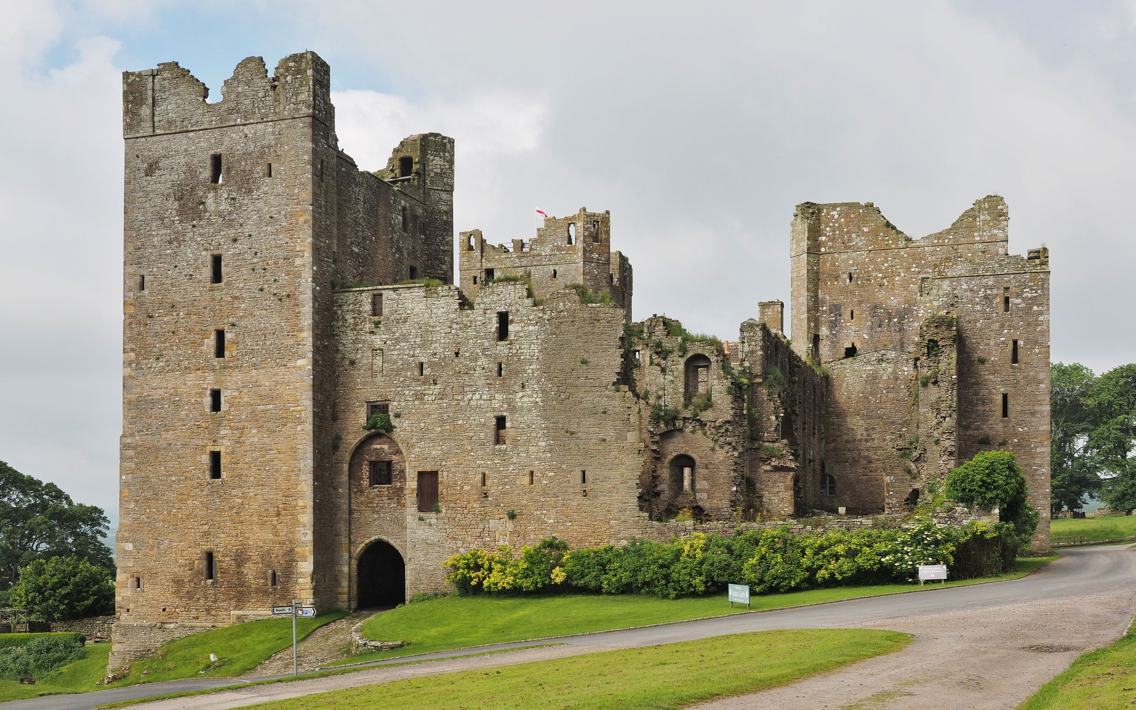 A historic castle with multiple towers and arched windows, surrounded by lush greenery and a paved road.