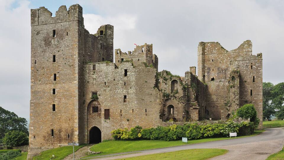 A historic castle with multiple towers and arched windows, surrounded by lush greenery and a paved road.