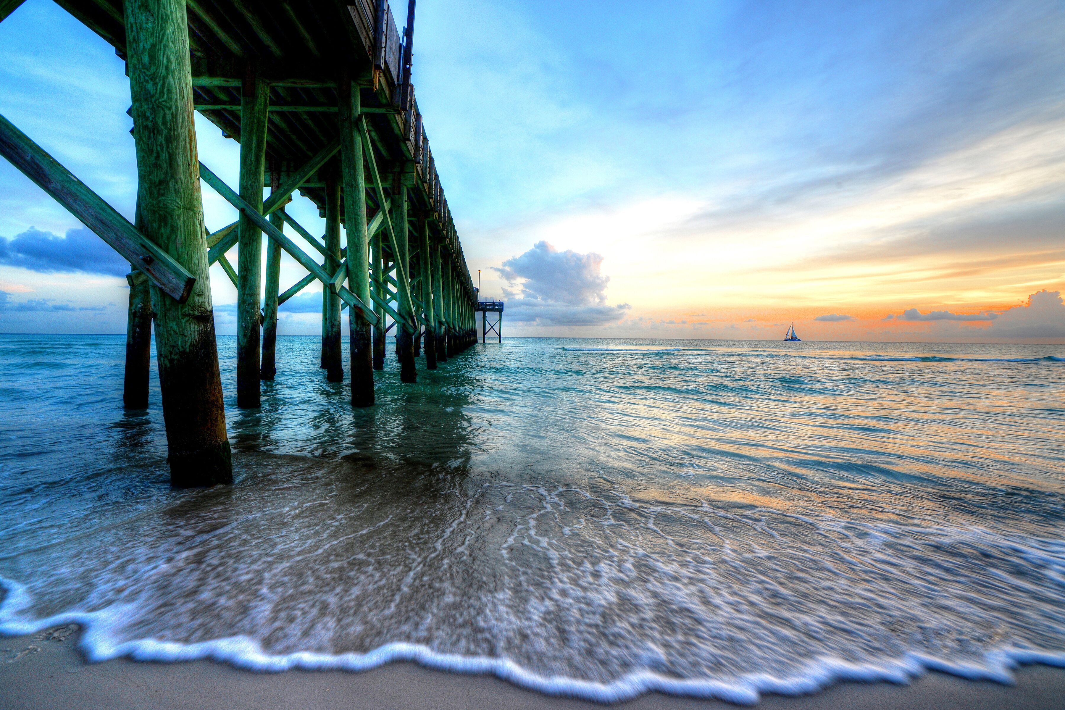 A pier extending into calm waters at sunset with a sailboat in the distance.
